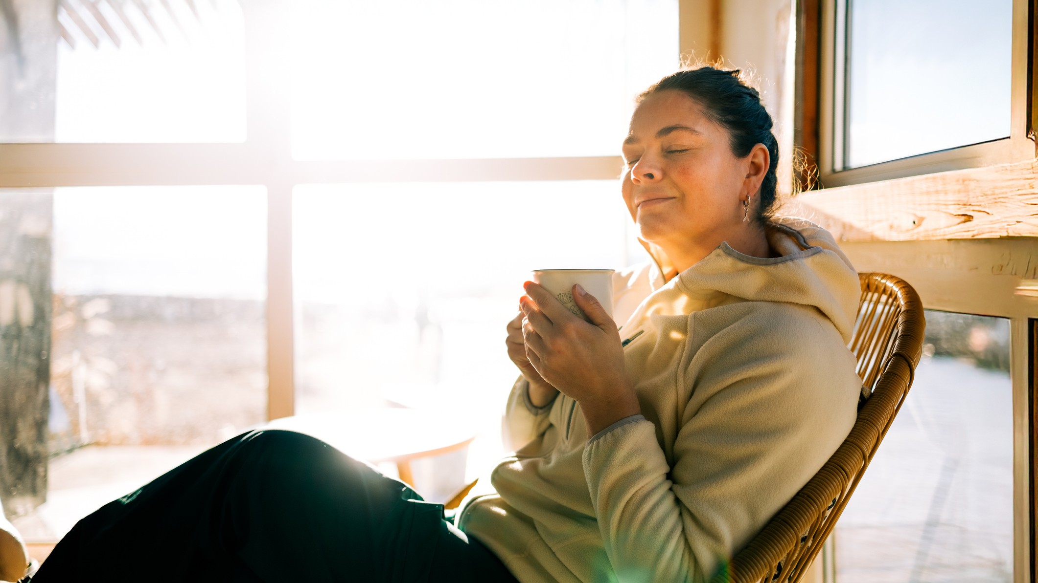 a woman relaxes in a chair, sipping coffee near windows