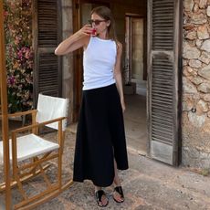 Danish fashion influencer and fashion industry creative Clara Dryhauge poses outside Mallorca hotel room wearing tort square sunglasses, a white tank top, black midi skirt, and flat black sandals