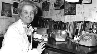 Italian scientist Rita Levi-Montalcini wearing a white gown sitting at a desk and holding a guinea pig's tail. Italy, 1950s