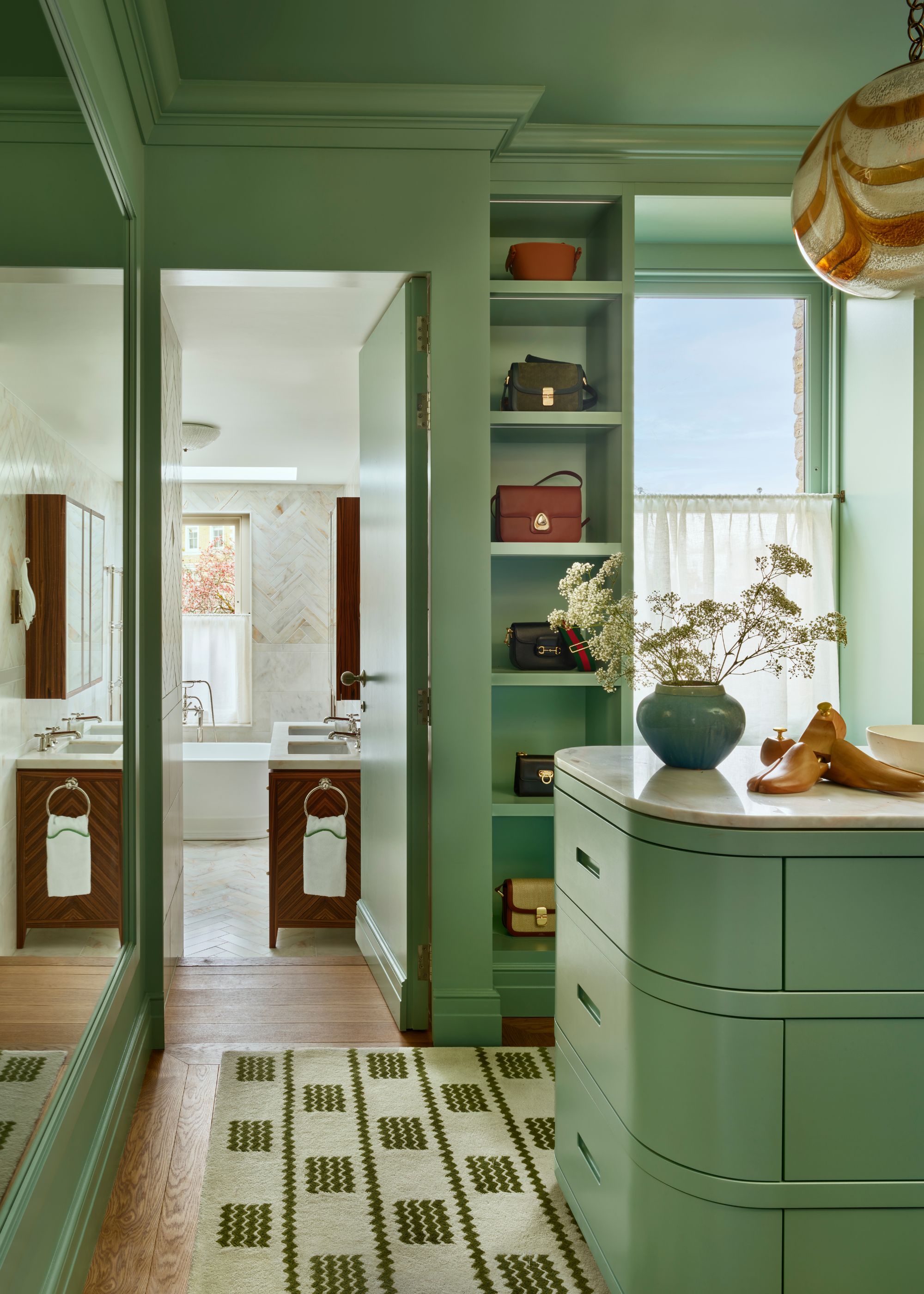 A dressing room with a pistachio green color with an island unit and built-in shelving. A patterned rug on the ground and a window with a cafe curtain.