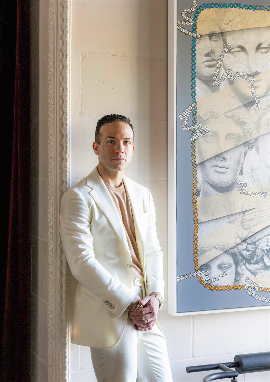 a man in a white jacket and pants standing against a wall and next to a large painting of Roman bust sculptures 