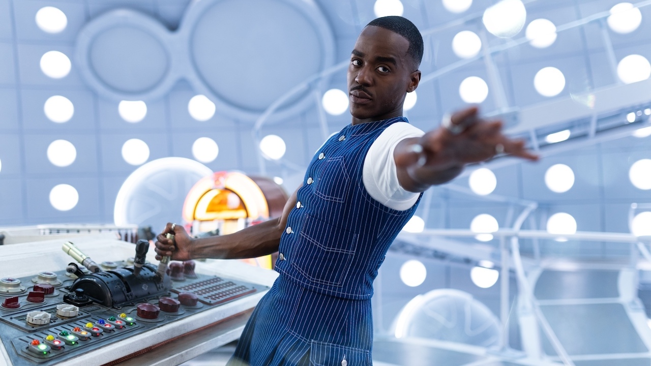Fifteenth Doctor wearing TARDIS-inspired blue vest and kilt, reaching his hand out while next to TARDIS console