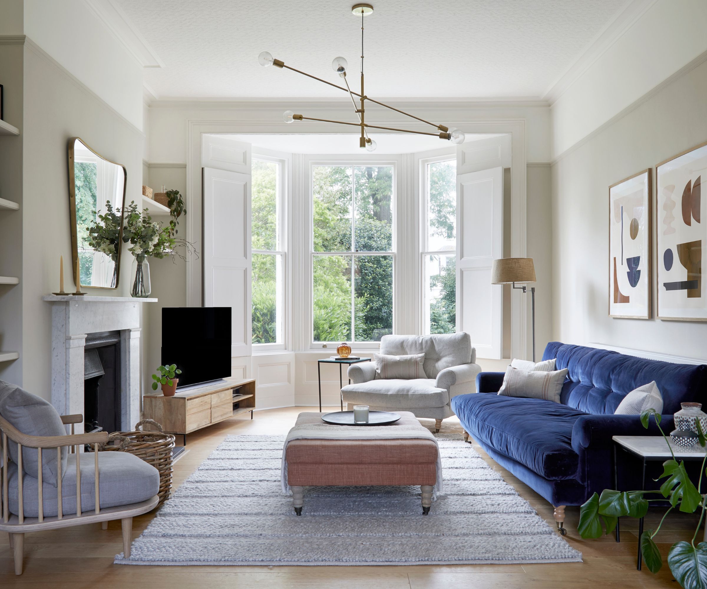 Living room with taupe walls, large bay window, grey rug at the centre, a pink ottoman on top, dark blue velvet sofa, and a large fireplace with a mirror above it