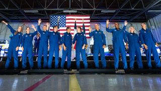 10 astronaut candidates stand in a row in a hangar, in front of an american flag and t-38 jets