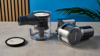 the empty dust basked of a modern-looking sleek shiny vacuum cleaner with light grey components on a carpet in front of a blue background