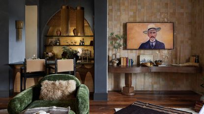 Modern Hacienda TV Room with grey walls, arched cabinetry bar with a gold background, green velvet armchair, dark wooden flooring, wooden built-in table, tiled wall with frame TV and books and plants
