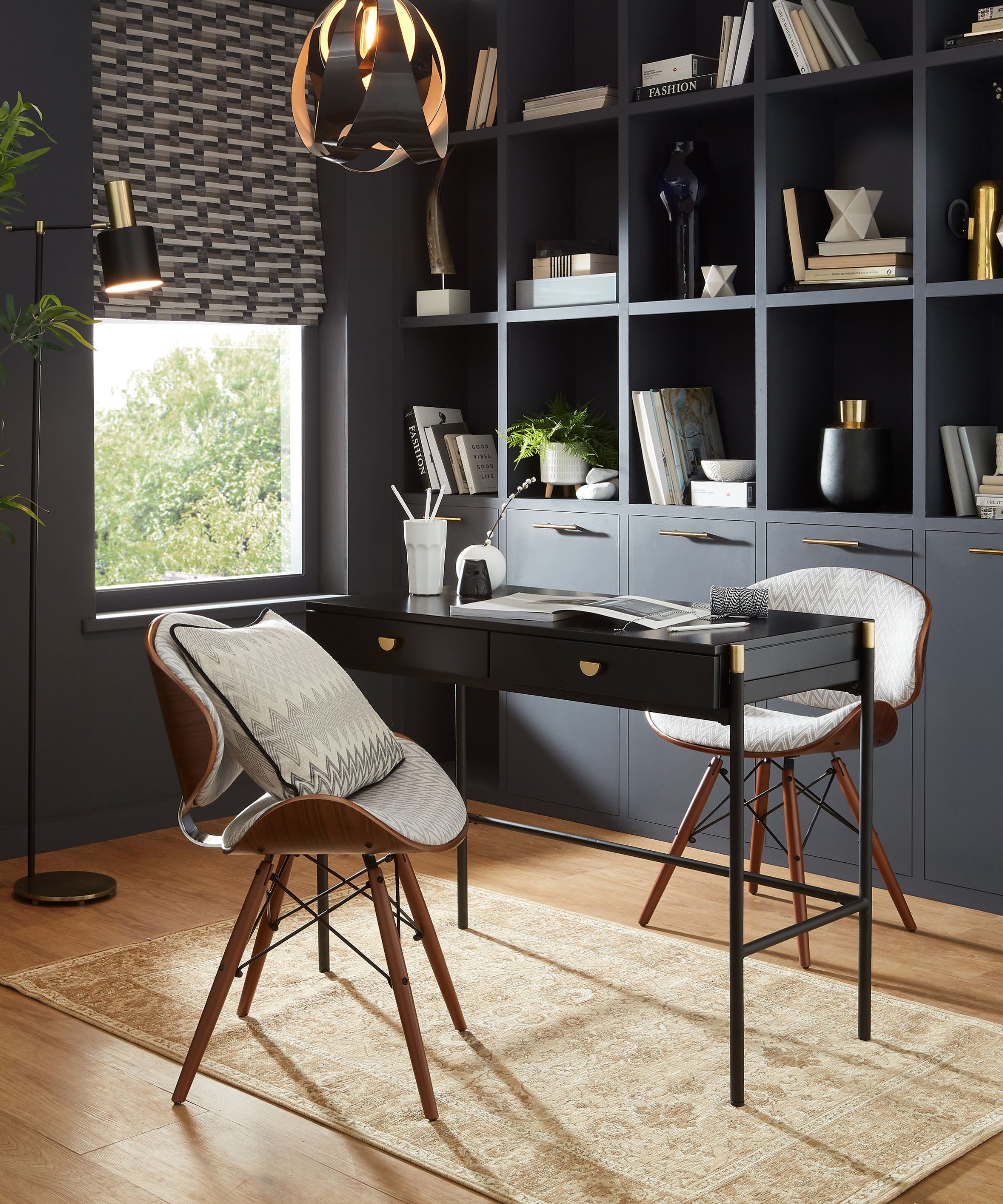 A stylish office with a black desk and two mid-century chairs sits in front of deep navy built-in shelves, layered with monochrome books and sculptural decor.