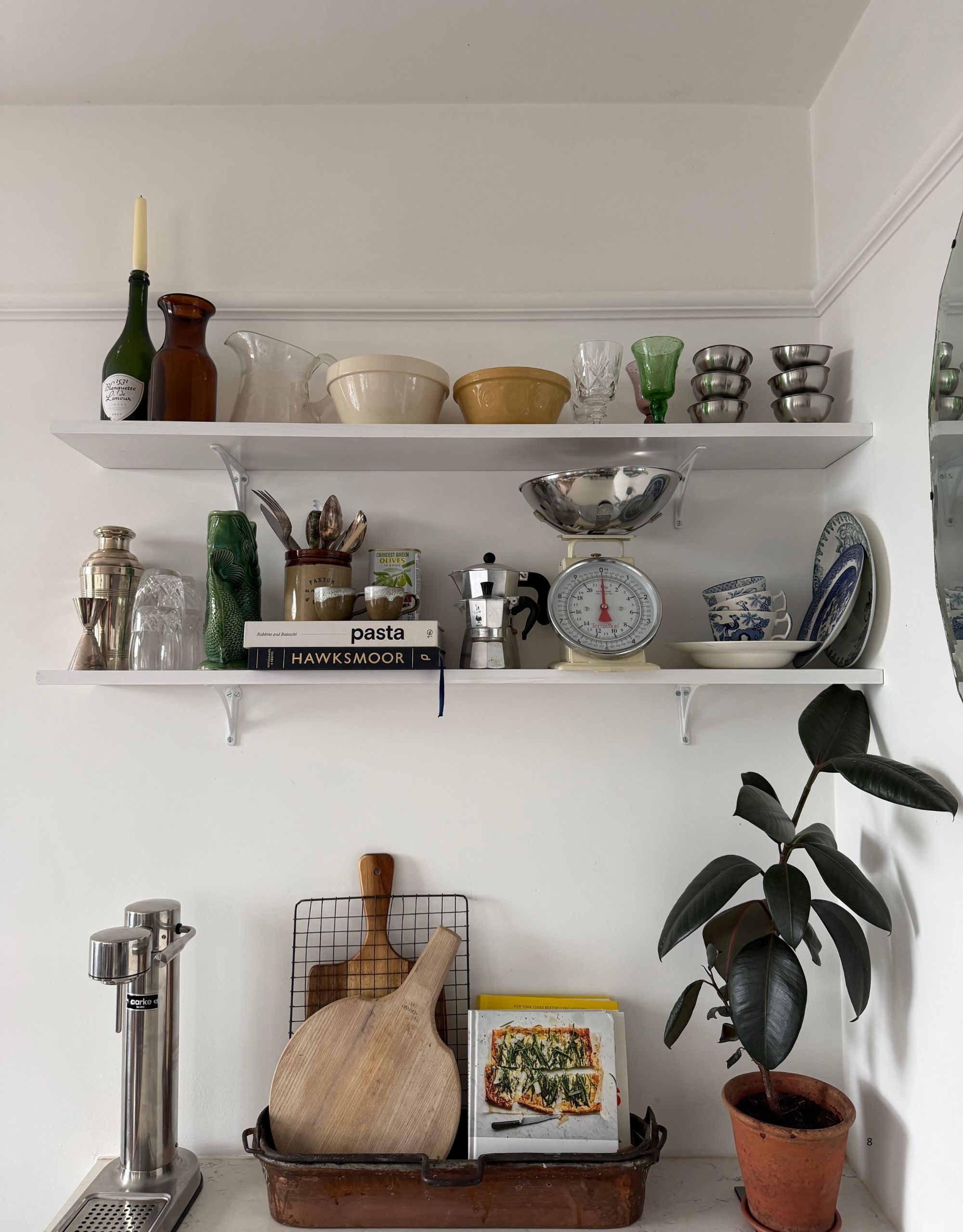 Image of a white wall with open shelves that are filled with different kitchen items. There are ceramic bowls, silver cups, silver coffee pots, blue and white china, and more.