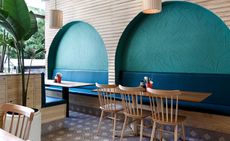Wood designed dining area with green and blue recessed arch in wall designs, hanging lights, long tables with a mix of leather sofa seating and wooden chairs 