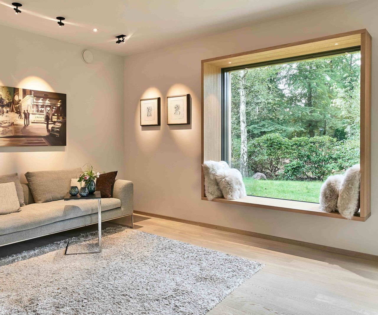 The living room of a timber-clad self build, built by Baufritz. The room features a large picture window with window seat overlooking woodland, and neutral walls with artwork illuminated from above. There is also low grey sofa and soft grey rug.