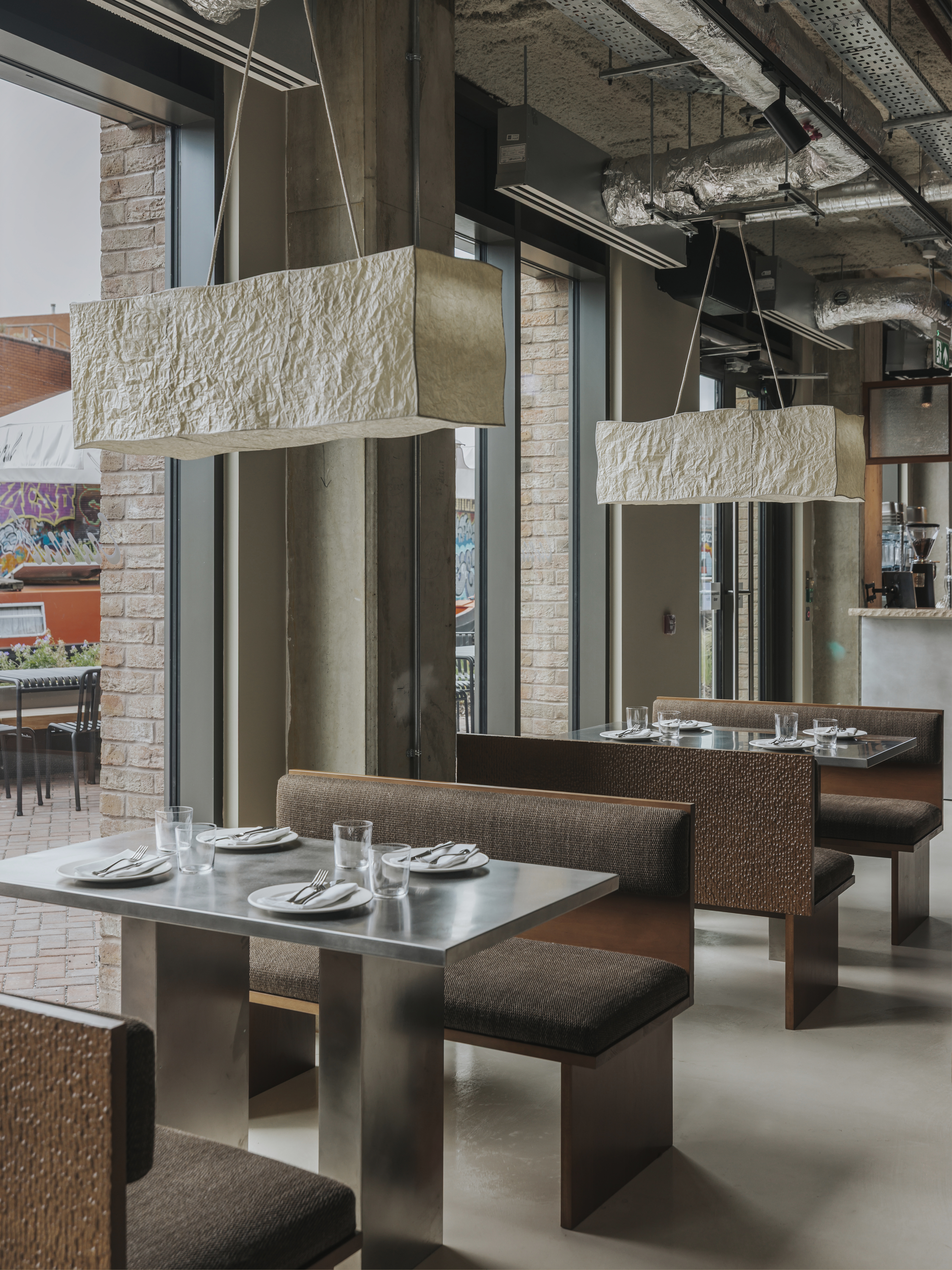 A restaurant with brutalist interiors features wooden upholstered banquettes with warm, brown fabric matching with matte chrome tables with a geometric shape.