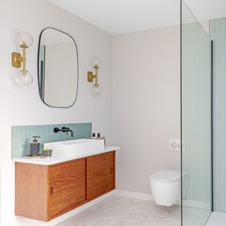 A modern bathroom with glass shower panel, white toilet fitted to wall, teak wood vanity and soft rectangular mirror