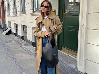 Fashion influencer Felicia Akerstrom poses on a sidewalk in Copenhagen wearing black oval sunglasses, a trench coat, white t-shirt, jeans, and black leather shoulder bag.