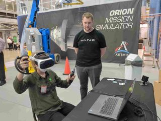 a person using a virtual reality headset and controllers while sitting in front of a laptop computer. a person in a t-shirt stands beside. in the backdrop is a big sign saying "orion mission simulator"