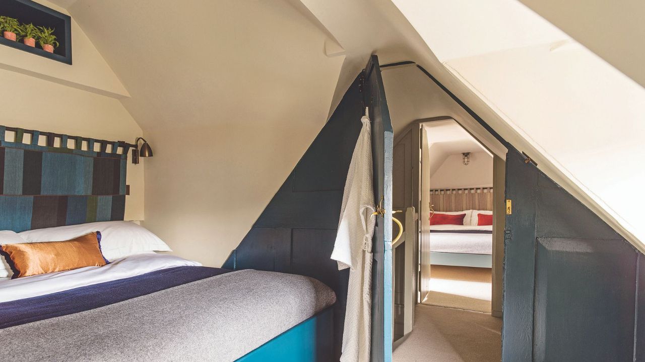 A white painted attic bedroom with dark teal feature wall, a patterned blue fabric headboard with a small shelf above with three potted plants and an open door to another bedroom behind