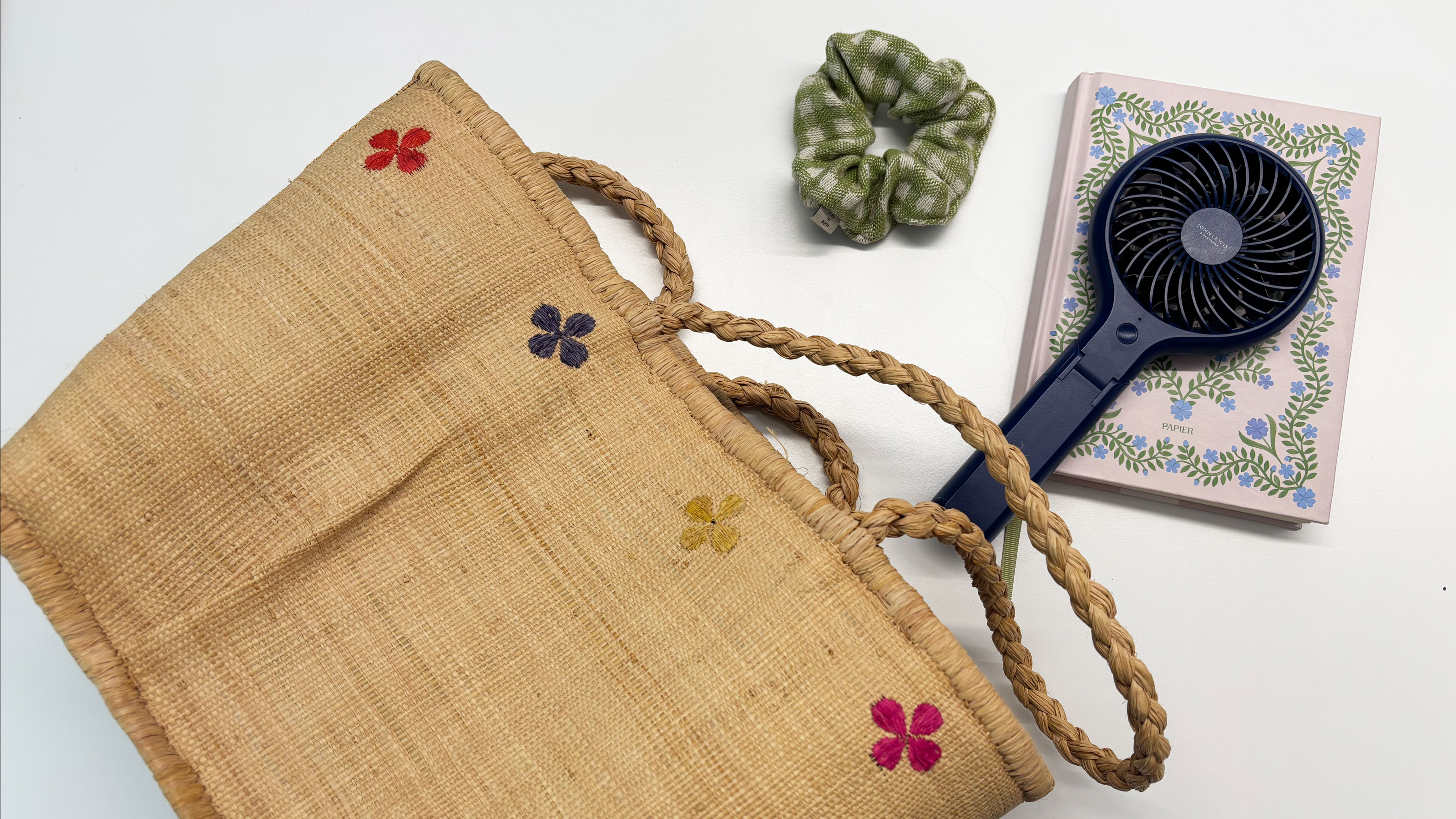 Straw bag on white desk with blue fan, pink notebook and green scrunchie