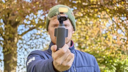 Man in beanie hat holding the DJI Osmo Pocket 4 vlogging camera with its magnetic fill light, an autumnal tree behind him