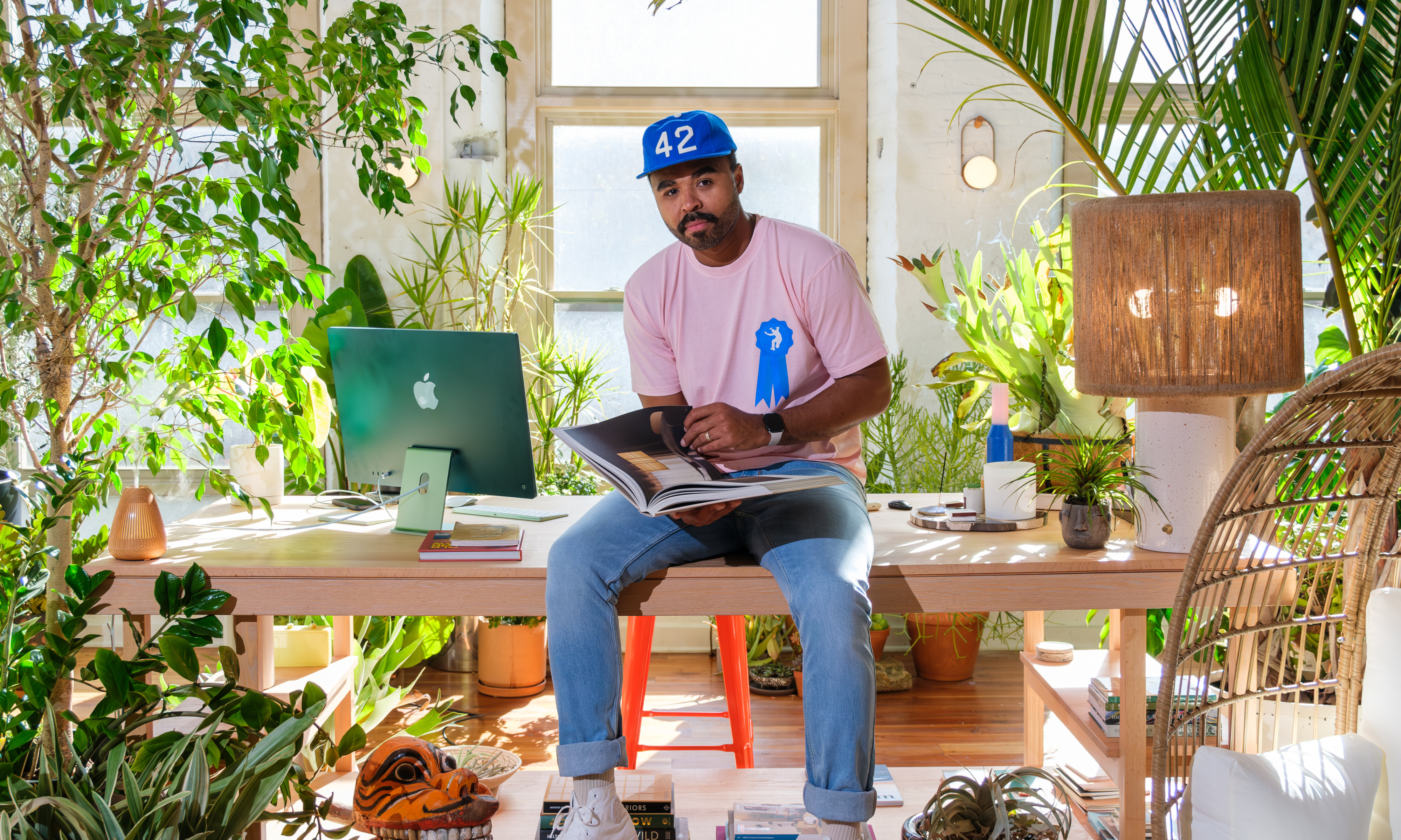 Man in jeans and pink t-shirt sat on office desk surrounded by large, topical indoor plants