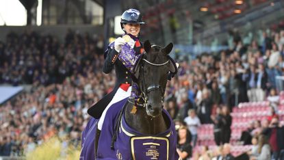Lottie Fry holds her medal aloft as she trots Glamourdale, who is wearing a purple winner's rug, towards the camera with the crowds in the stands behind