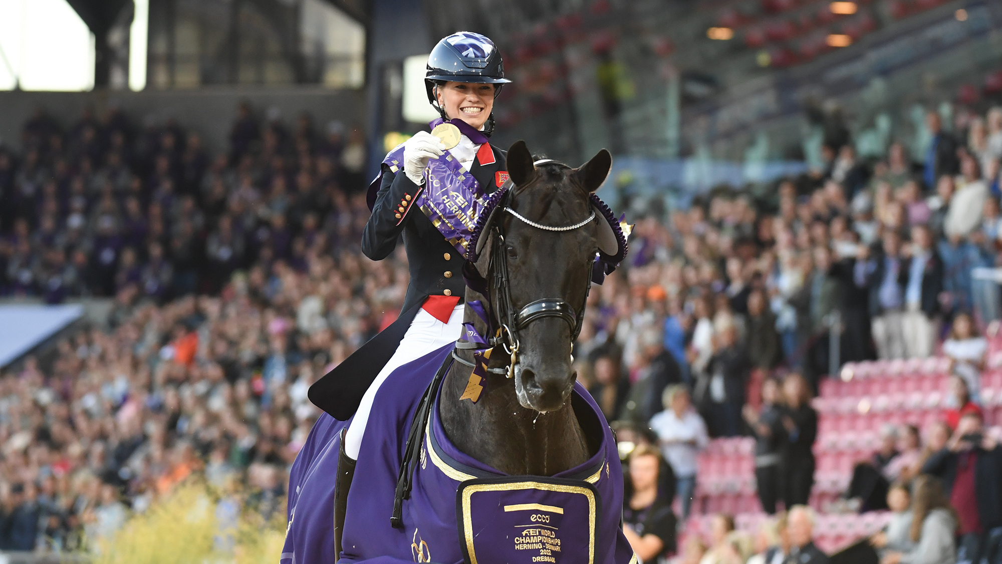 Lottie Fry holds her medal aloft as she trots Glamourdale, who is wearing a purple winner's rug, towards the camera with the crowds in the stands behind