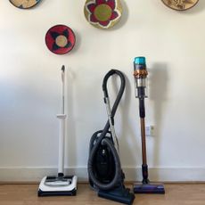 A gtech upright, miele cylinder, and dyson stick vacuum standing next to each other in front of a white wall