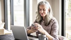 An older woman looks at her smartphone while sitting at the kitchen table, her laptop open in front of her.