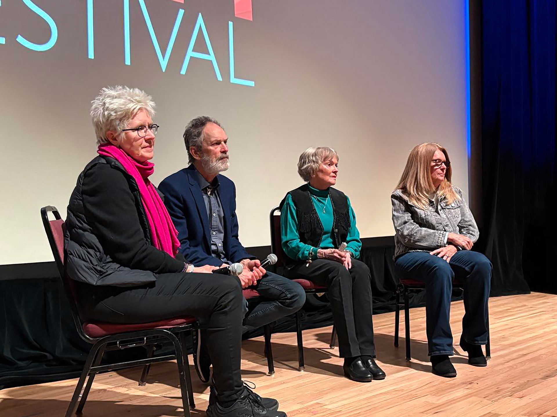 three women and a man sit on a stage with a movie screen behind them