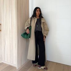 Stylist Monikh Dale wearing a fall outfit including a classic Toteme trench coat, grey t-shirt, black pants, green The Row tote bag, and Adidas SL72 sneakers.