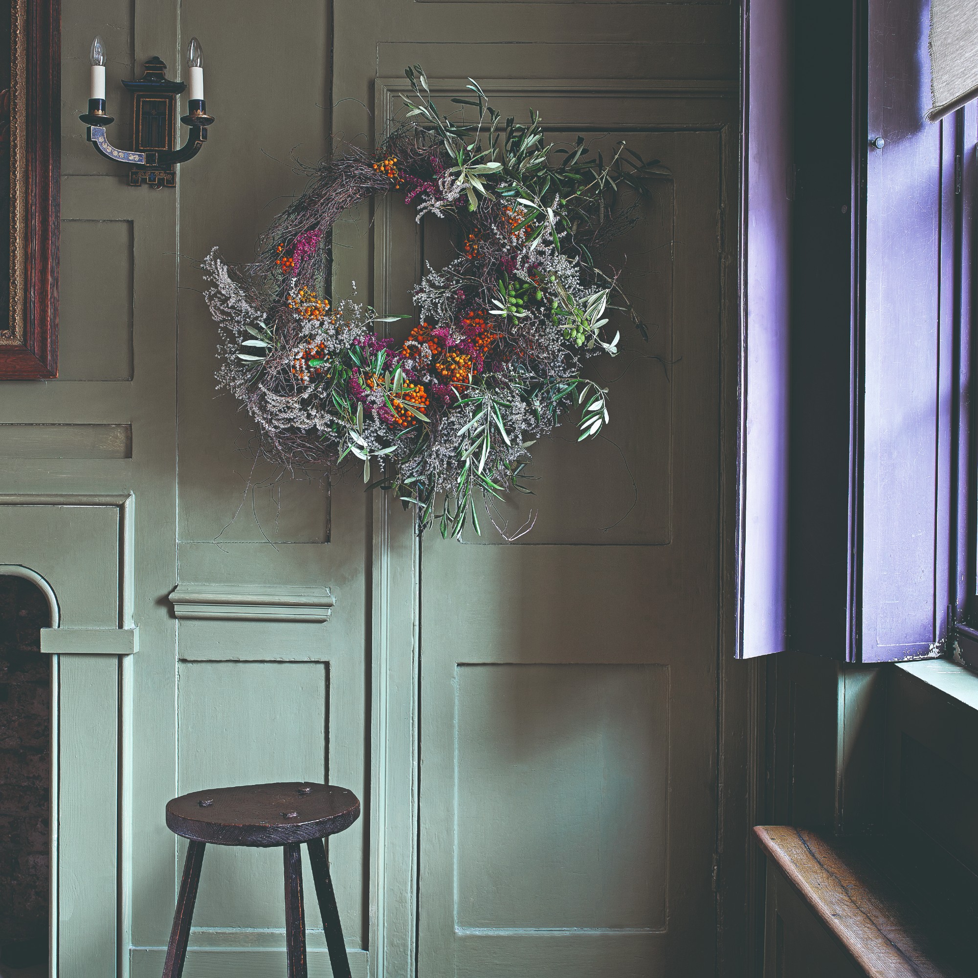 A sage green-painted room with purple window shutters decorated with a large Christmas wreath