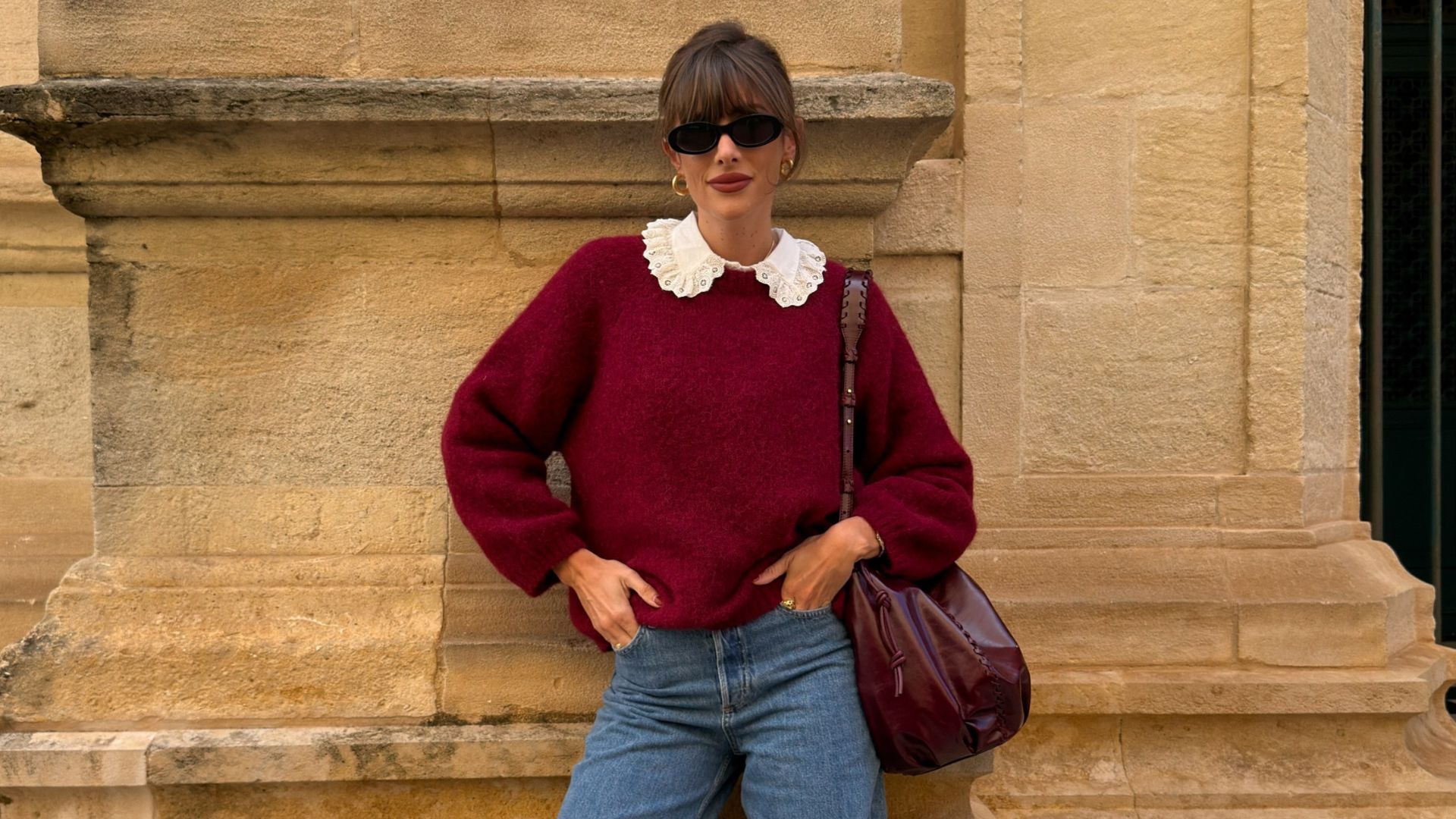 Influencer @juliesfi poses for a photo wearing a burgundy knit with blue jeans, brown loafers and a burgundy bag.