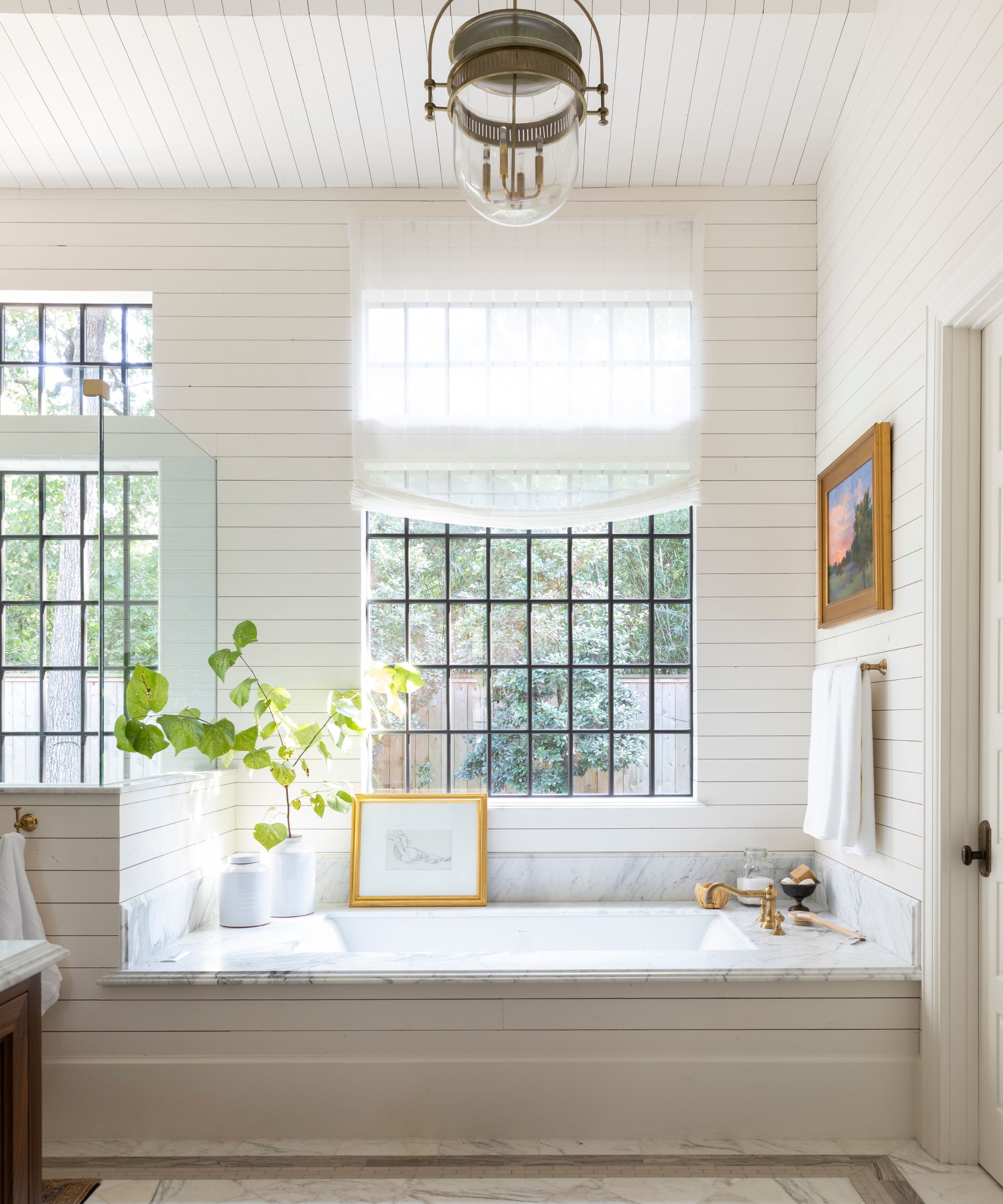 Bathroom with shiplap walls painted in Cloud White by Benjamin Moore, large crittall windows, and a marble bathtub with a gold tap and vintage art around it