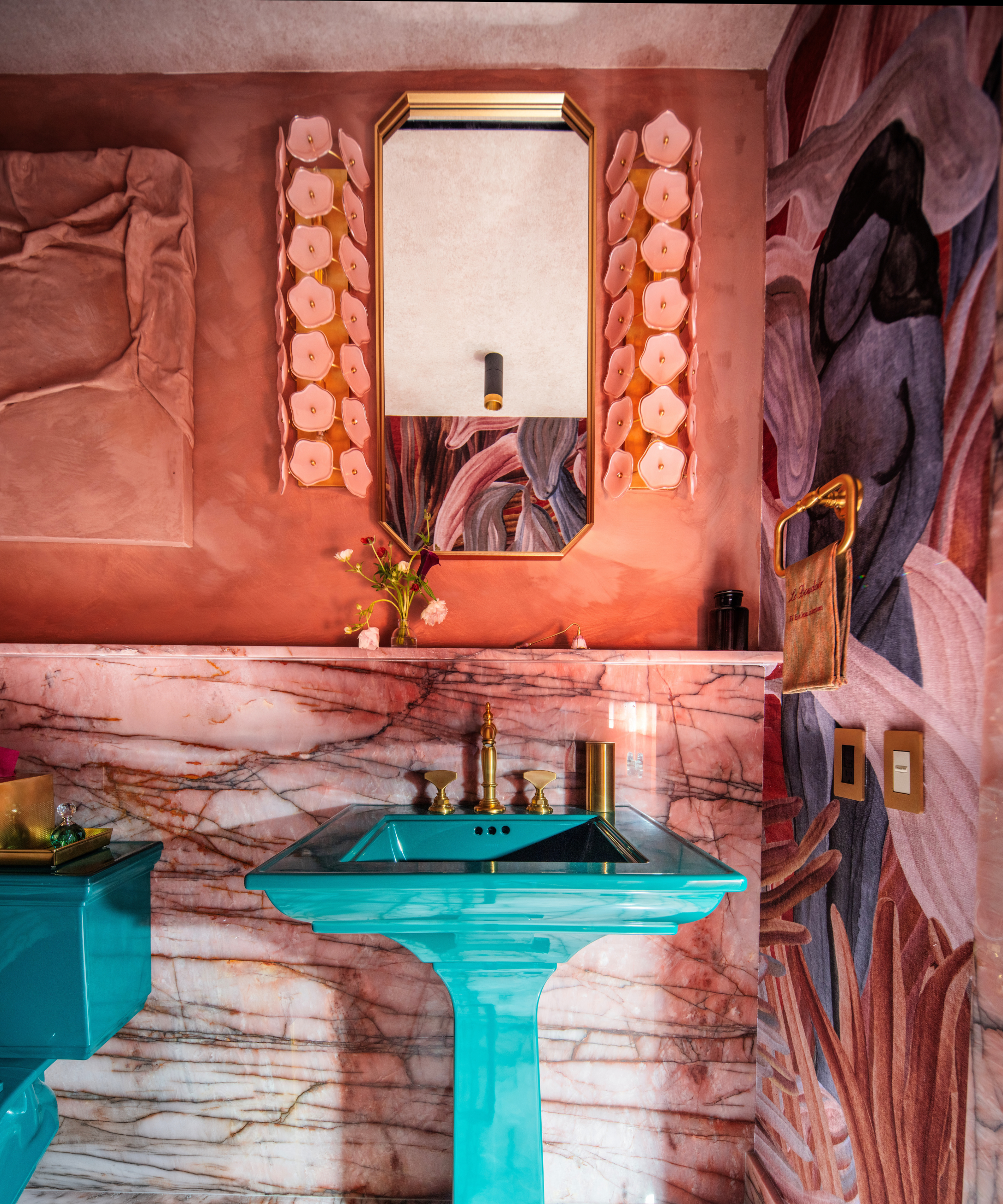 Artful powder room featuring a teal Kohler toilet and sink area juxtaposed with veiny red walls, a red-and-purple wallpaper textile, and pink floral wall sconces flanking the angular vanity mirror