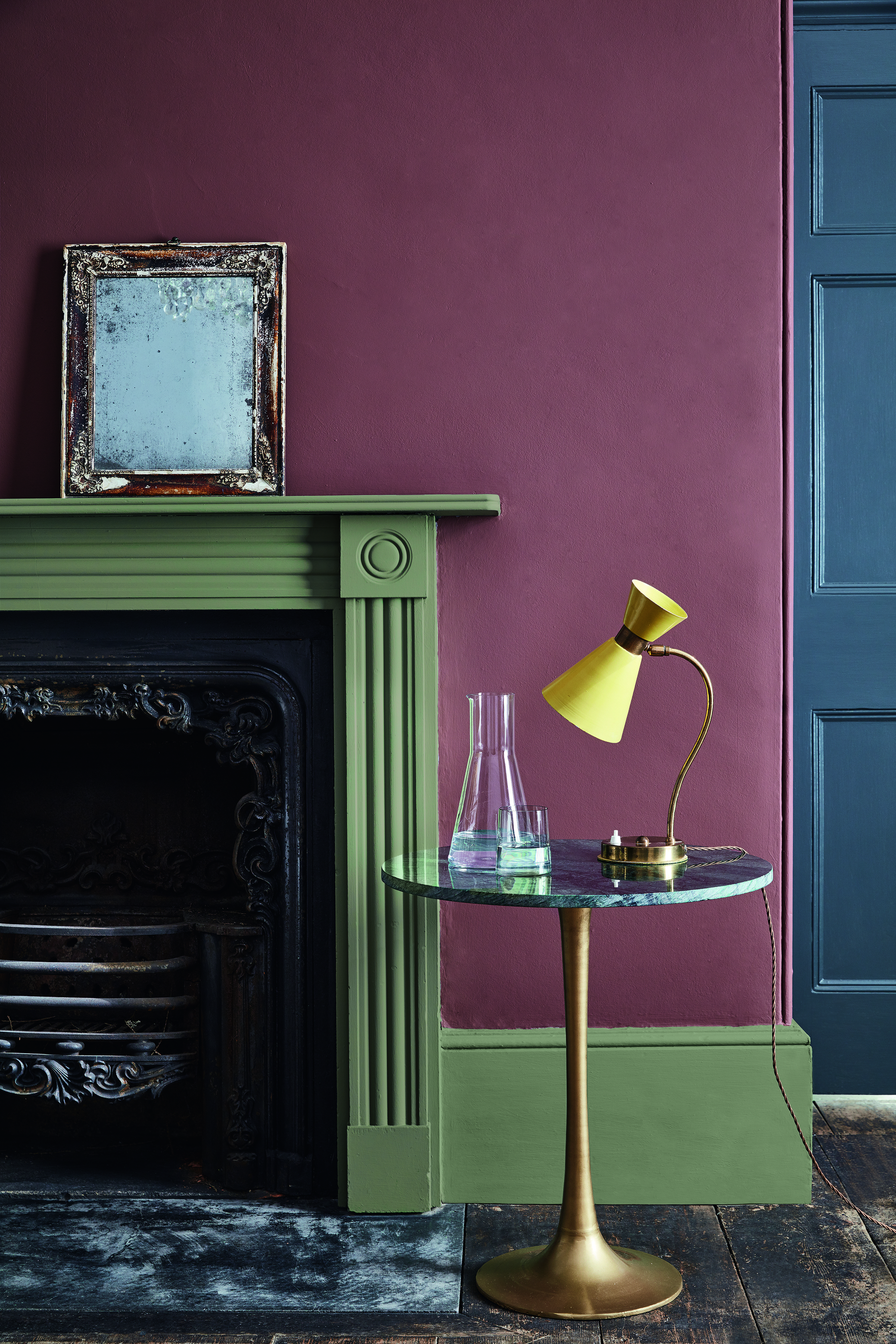 Close-up shot of a burgundy-painted wall and a fireplace painted in an olive green trim. There is a small marble side table with a glass carafe and a yellow lamp.
