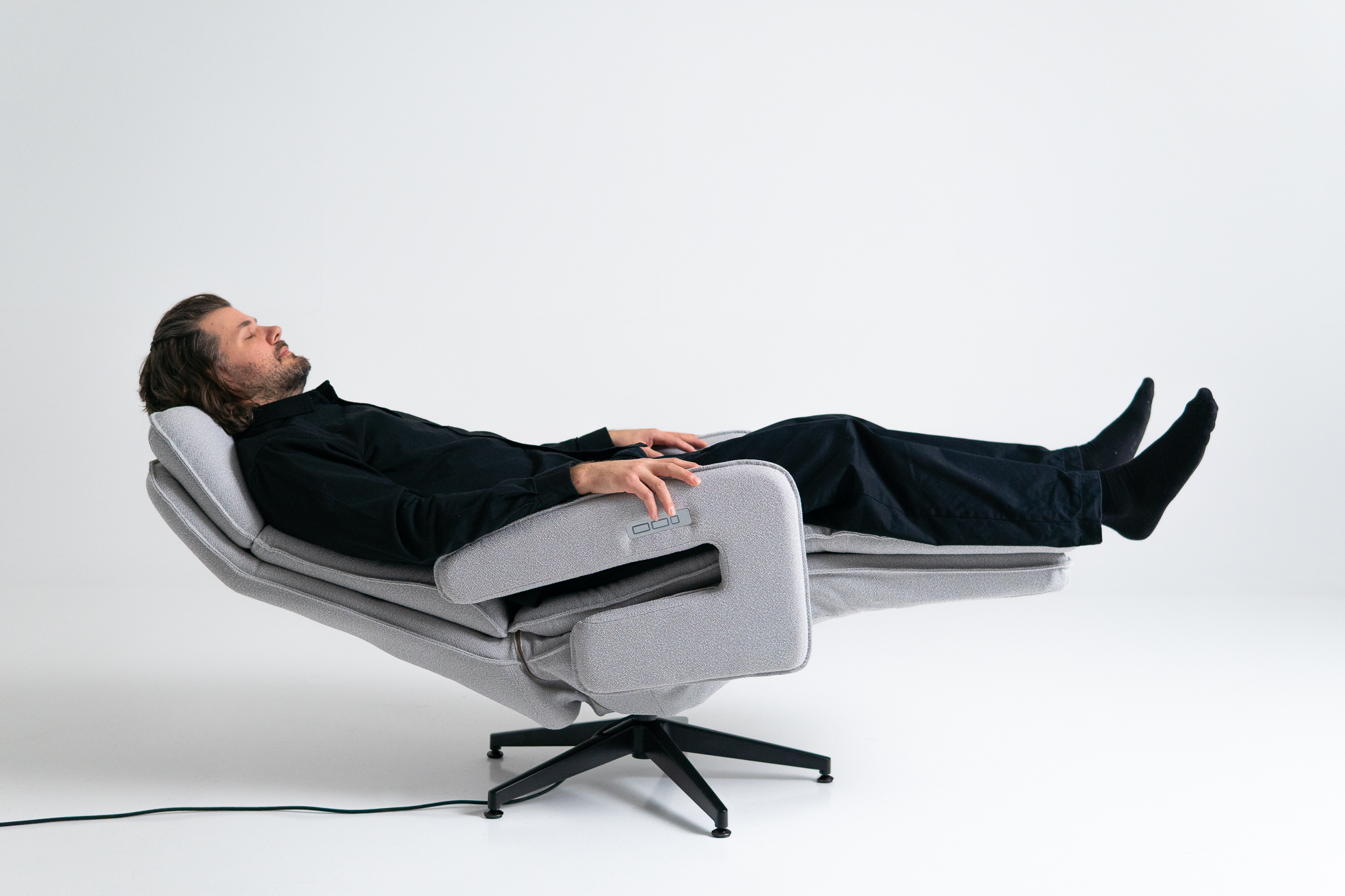 Studio product shot of a man fully reclined in the off-white Neurosonic Serene chair with his eyes closed, one hand resting on the armrest control panel.