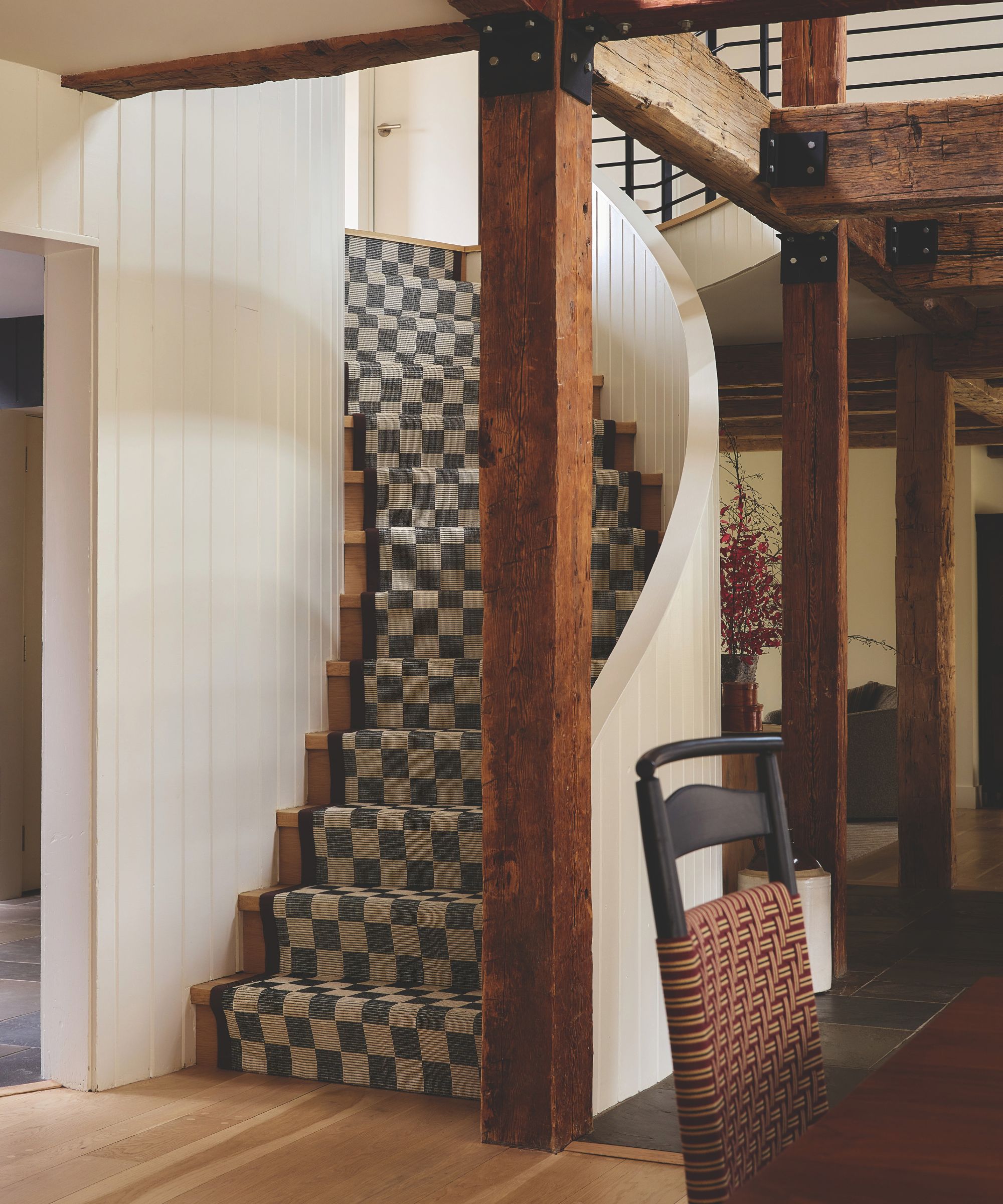 Curved wooden staircase featuring a black-and-beige checkered runner, framed by natural timber columns and white paneled walls.