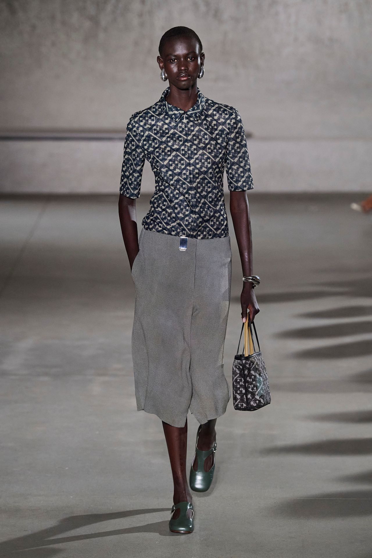 A model at the Tory Burch show wearing a patterned short-sleeve shirt, gray culotte shorts, and gray-silver shoes at the S/S 24 show.