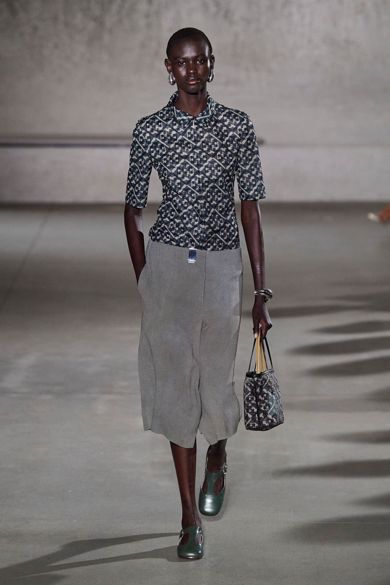 A model at the Tory Burch show wearing a patterned short-sleeve shirt, gray culotte shorts, and gray-silver shoes at the S/S 24 show.