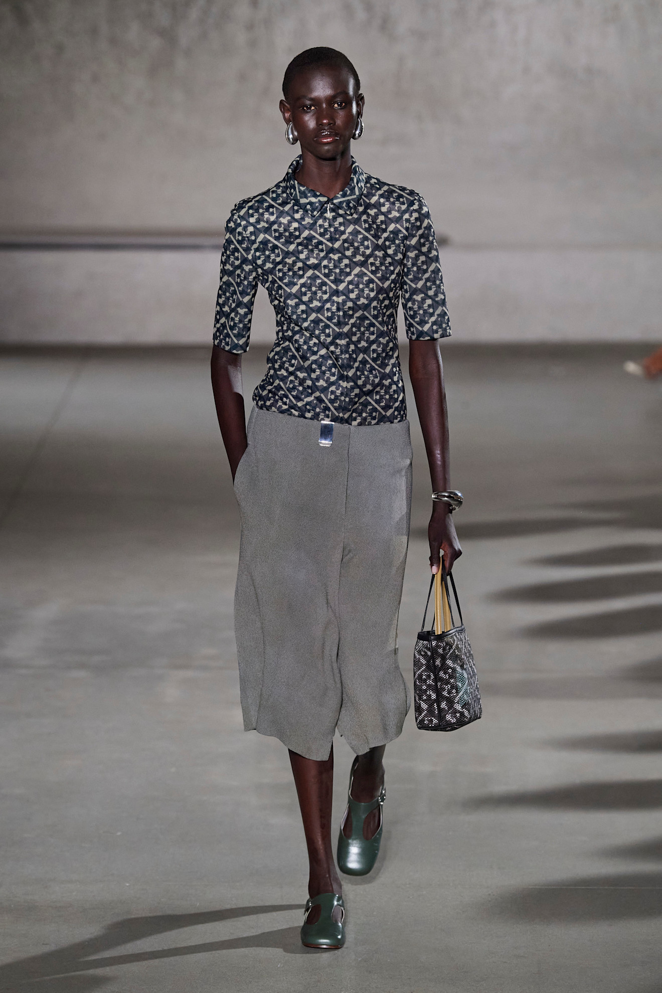 A model at the Tory Burch show wearing a patterned short-sleeve shirt, gray culotte shorts, and gray-silver shoes at the S/S 24 show.