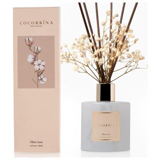 A white glass reed diffuser with gold detailing, brown reeds and white flowers, next to a pink packaging box with flower image