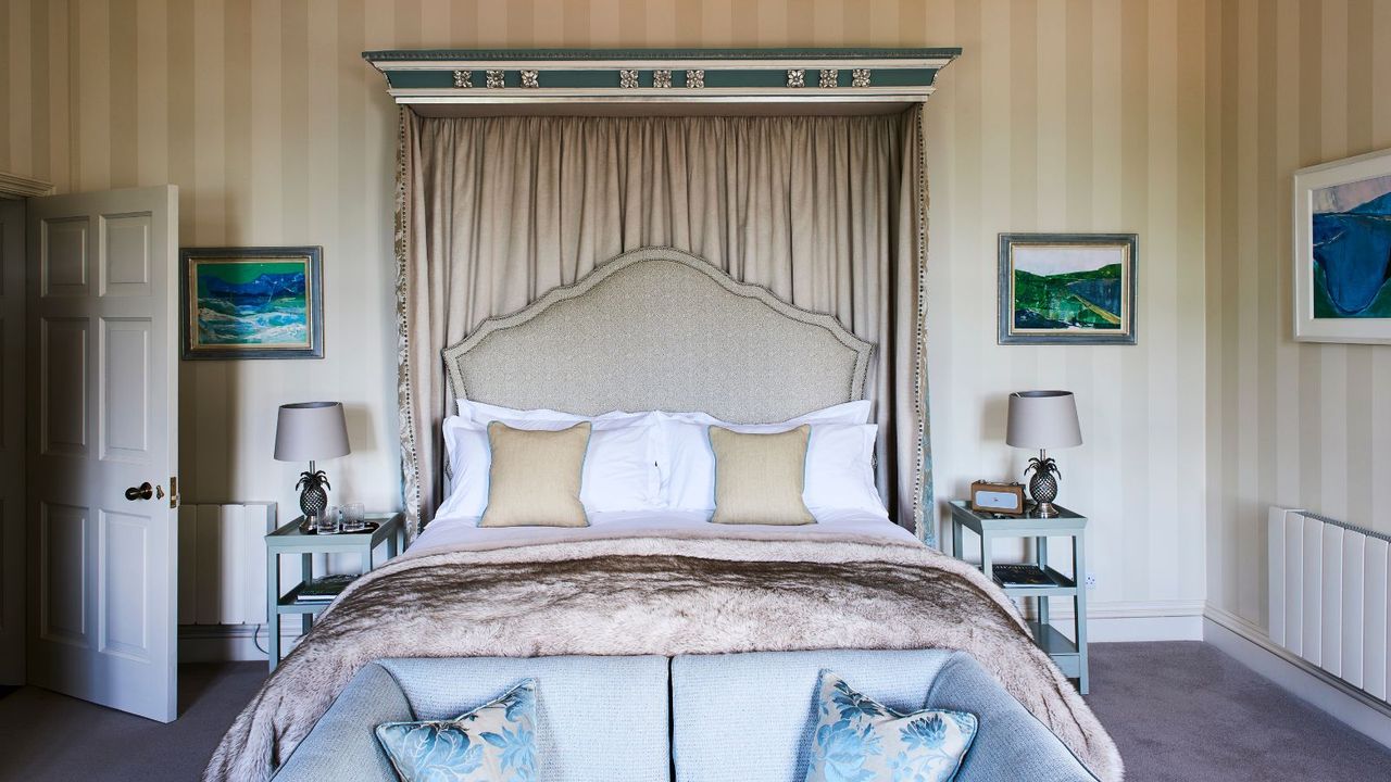 Bedroom with large double canopy bed and cream colored striped wallpaper. At the end of the bed is a pale blue sofa with matching pillows, and to either side of the bed are pale blue bedside tables with matching pineapple lamps.