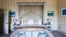 Bedroom with large double canopy bed and cream colored striped wallpaper. At the end of the bed is a pale blue sofa with matching pillows, and to either side of the bed are pale blue bedside tables with matching pineapple lamps.
