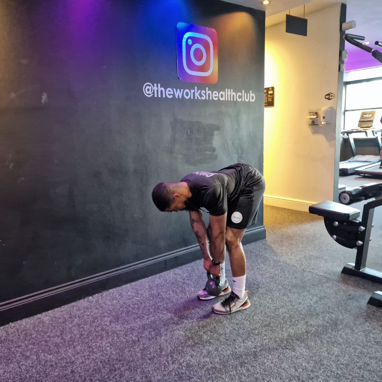 Renel Scarlett performs a kettlebell deadlift at The Works Health Club gym. He is bent over with a flat back, hinging at the hips with his arms hanging down and grabbing a kettlebell on the floor. Behind him we see empty treadmills.