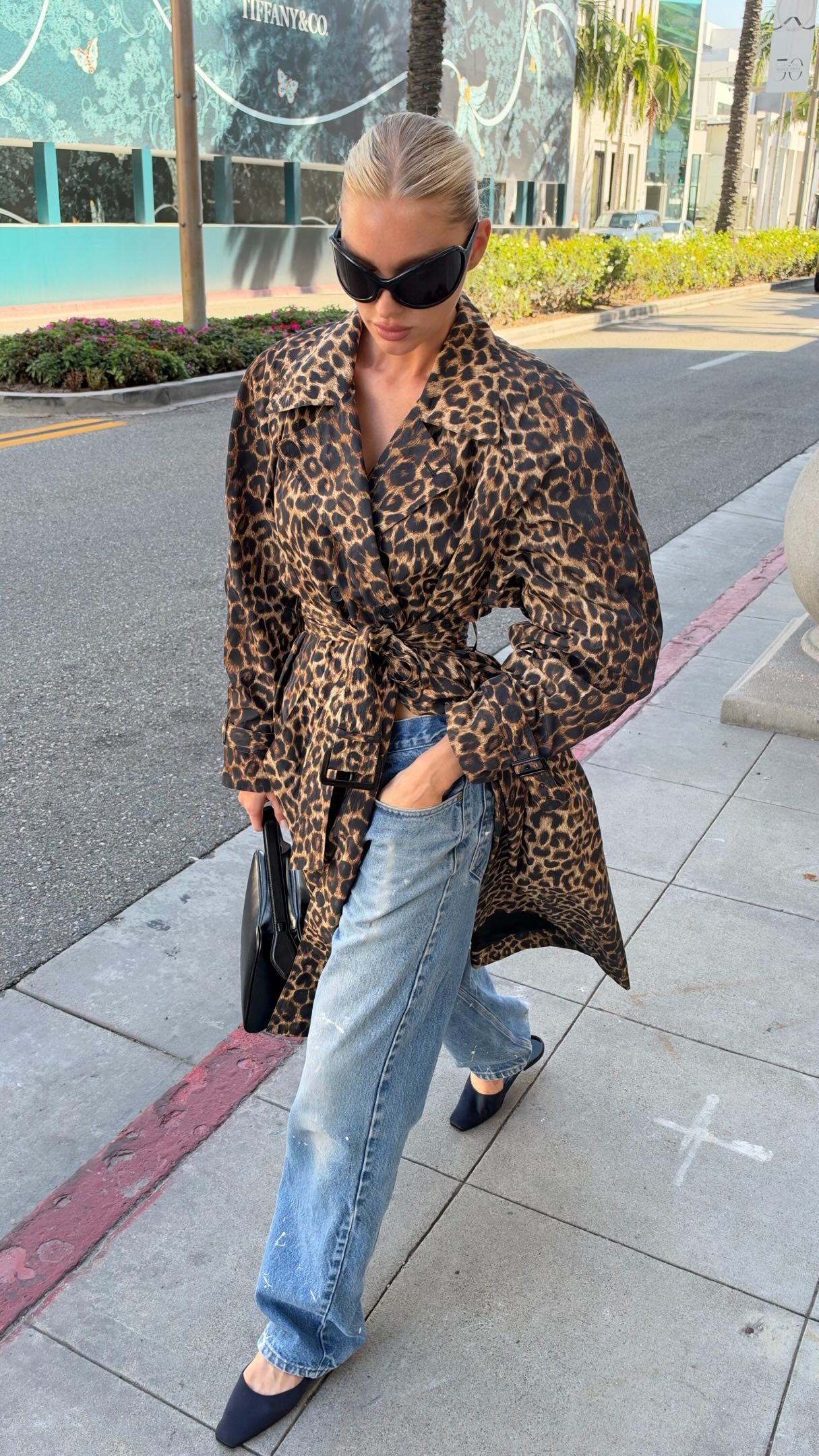 Elsa Hosk walks down the street wearing a leopard-print trench coat with relaxed, slightly distressed jeans and navy satin mules.