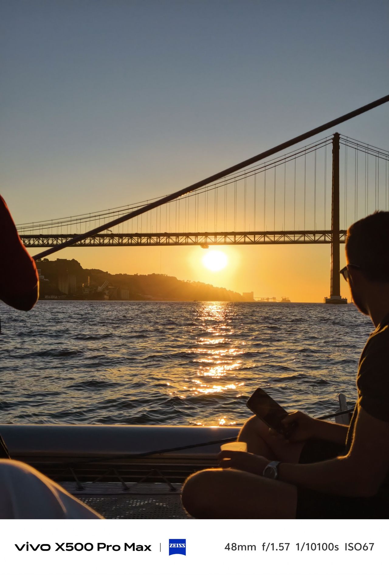 Sunset beneath the 25 de Abril Bridge, with golden light reflecting across the Tagus and a silhouetted passenger in the foreground.
