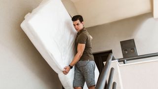 Man in brown shirt and grey shorts carries a white mattress down a flight of stairs
