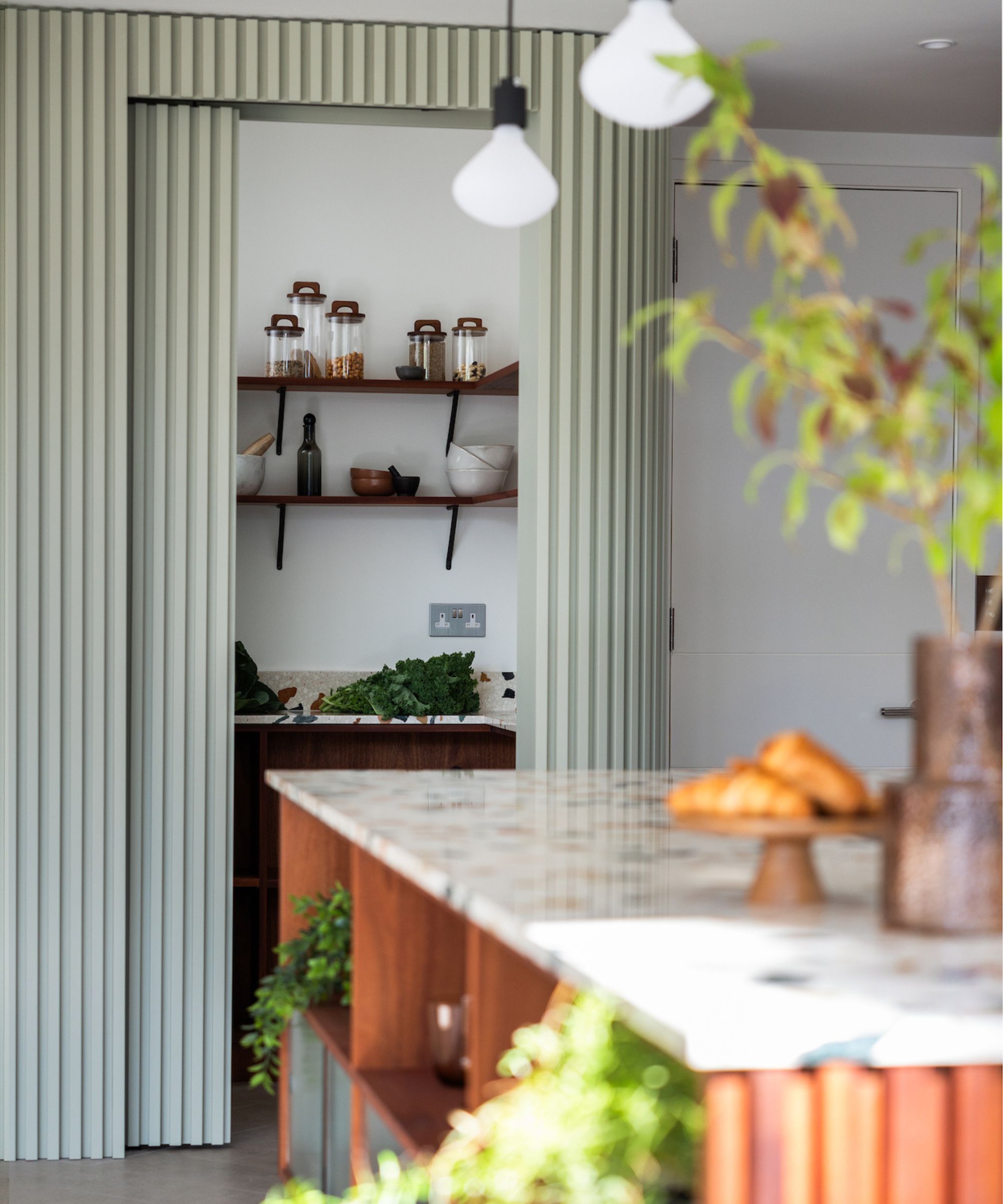 A sleek utility space concealed behind pale green slatted folding doors, blending beautifully with the modern kitchen design.