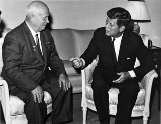 Black and white photo of two men sitting in chairs wearing dark suits and ties. The man on the left is bald.