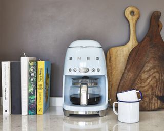 SMEG blue drip filter coffee maker on kitchen worktop with white mugs, wooden chopping board decor and assortment of hardback cookery books