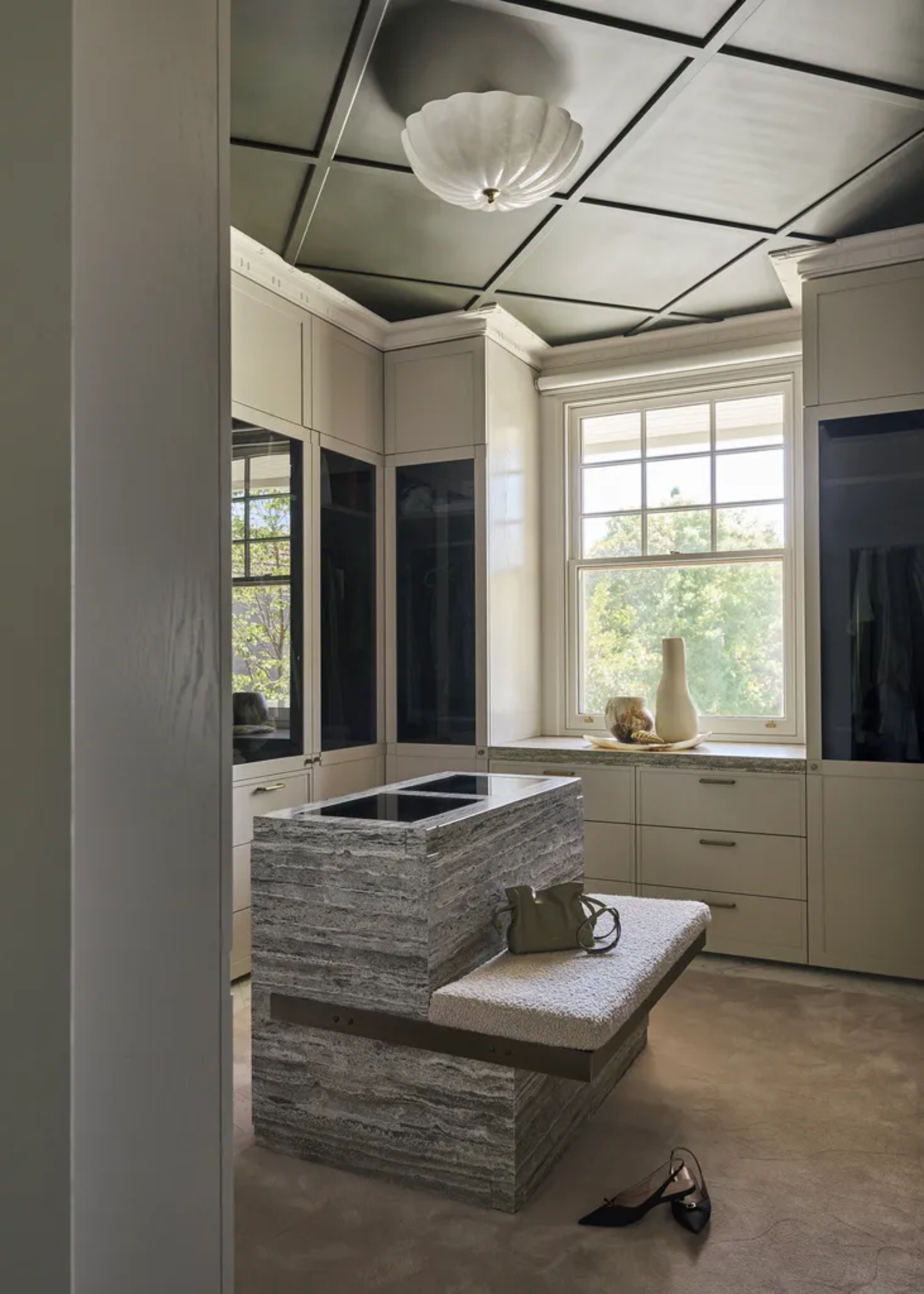 A walk-in wardrobe with white cupboard with tinted glass windows, a carpeted floor, and a dressing island with a bench holding a sage green bag above a pair of shoes