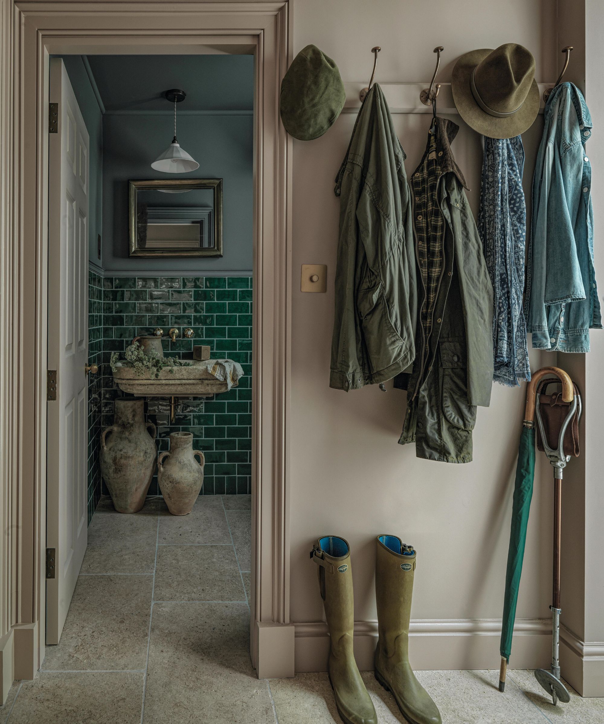a dusty lavender entryway with hooks for coats and boots on the floor, a view through to a dark blue bathroom with glossy green tiles
